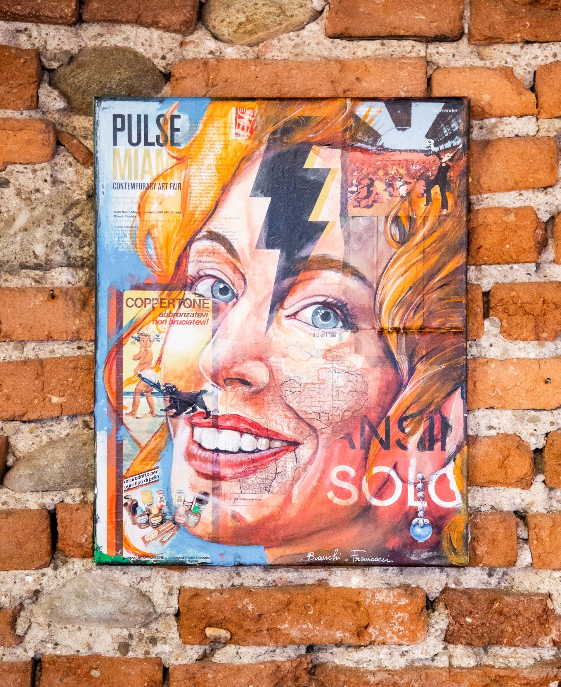 Opera a tecnica mista raffigurante il volto di una persona con un fulmine sovrapposto, esposta su un rustico muro di mattoni.