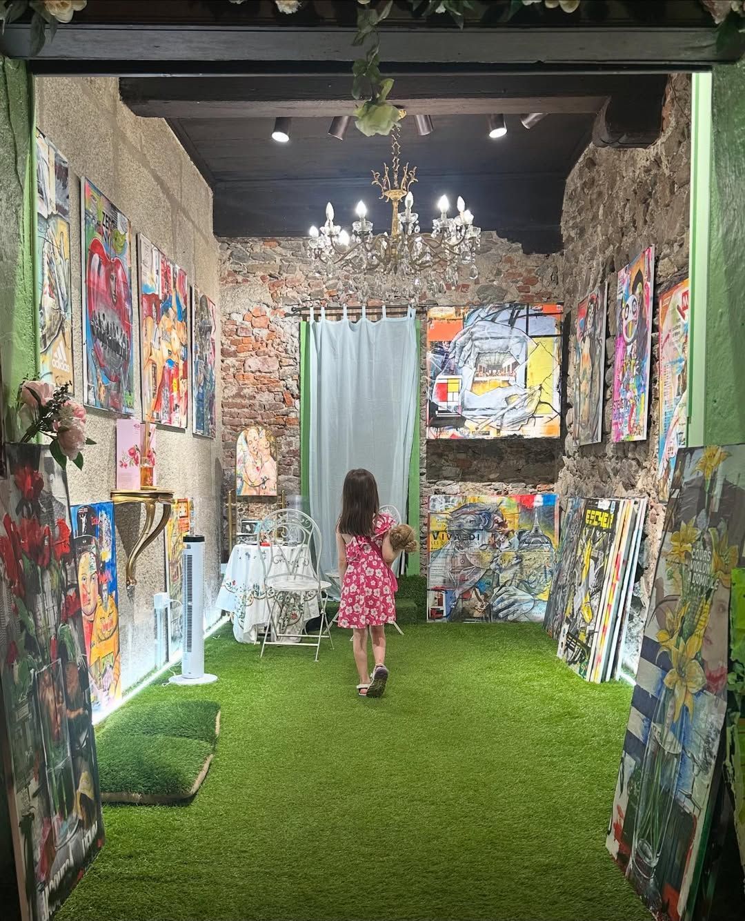 Un bambino cammina in una galleria al coperto con pavimento in erba, pareti in mattoni a vista e opere d'arte illuminate da un lampadario.