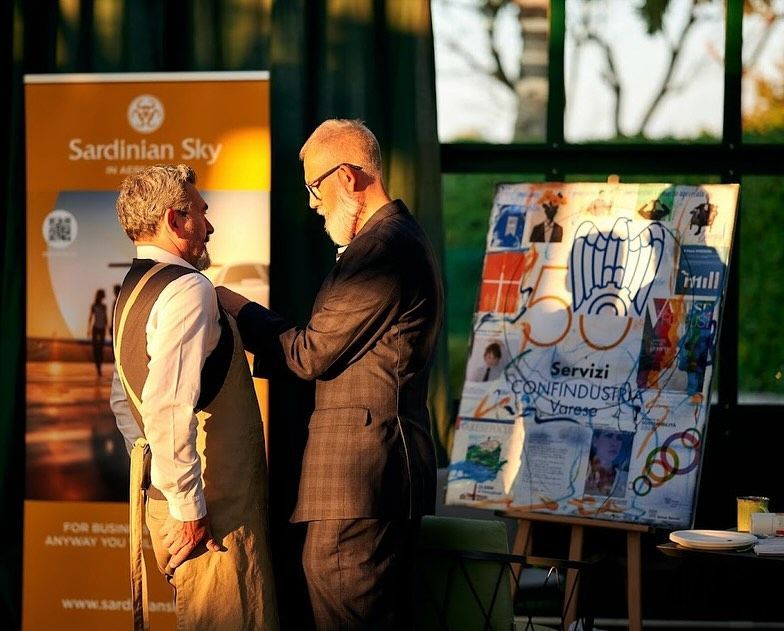In un ambiente interno illuminato dal sole, con striscioni e opere d'arte dell'evento nelle vicinanze, una persona appunta un badge a un'altra.