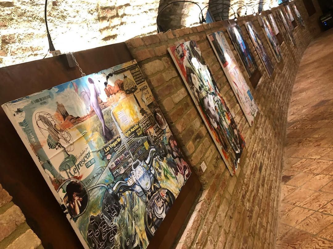 Una fila di opere d'arte astratte incorniciate è esposta su una parete di mattoni rustici all'interno di uno spazio espositivo scarsamente illuminato.