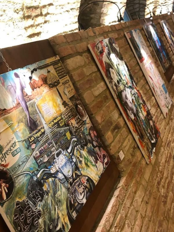 Una serie di opere d'arte realizzate con tecniche miste e appese a una parete di mattoni a vista, incentrate su un collage a tema motociclistico.