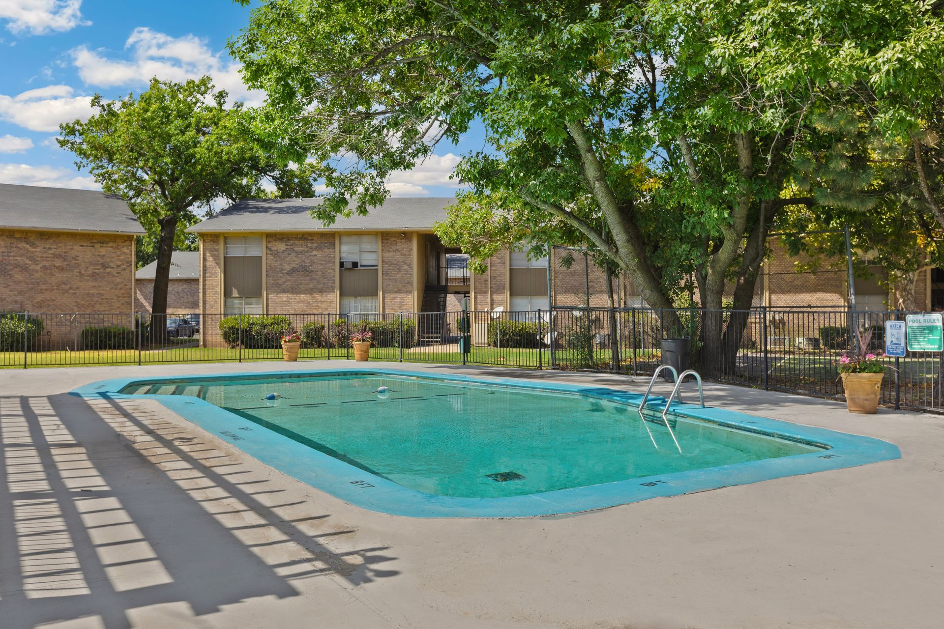 A swimming pool enclosed with fence, next to a building and trees.