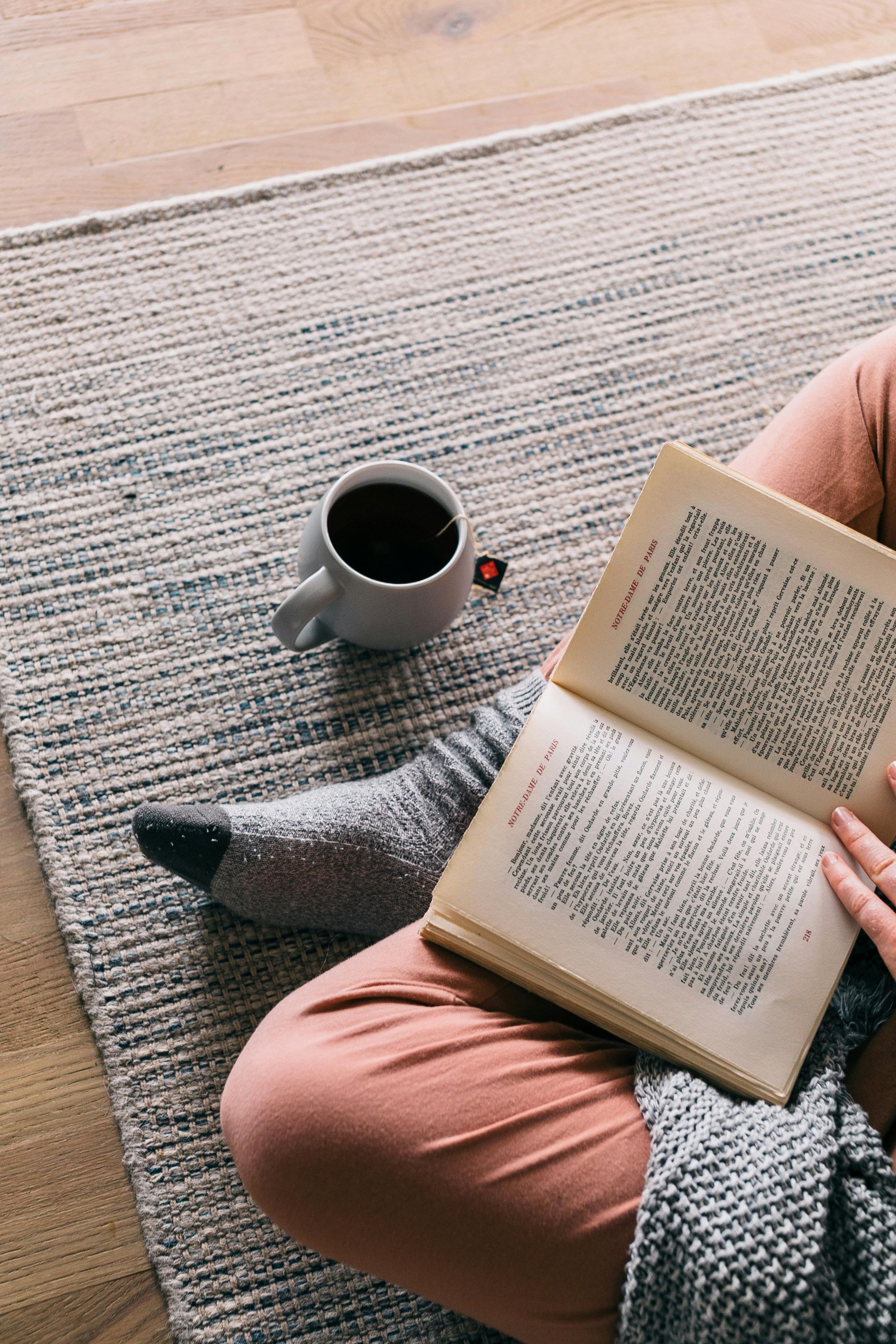 Person reading a book with a coffee cup on a rug, wearing socks and pink pants.
