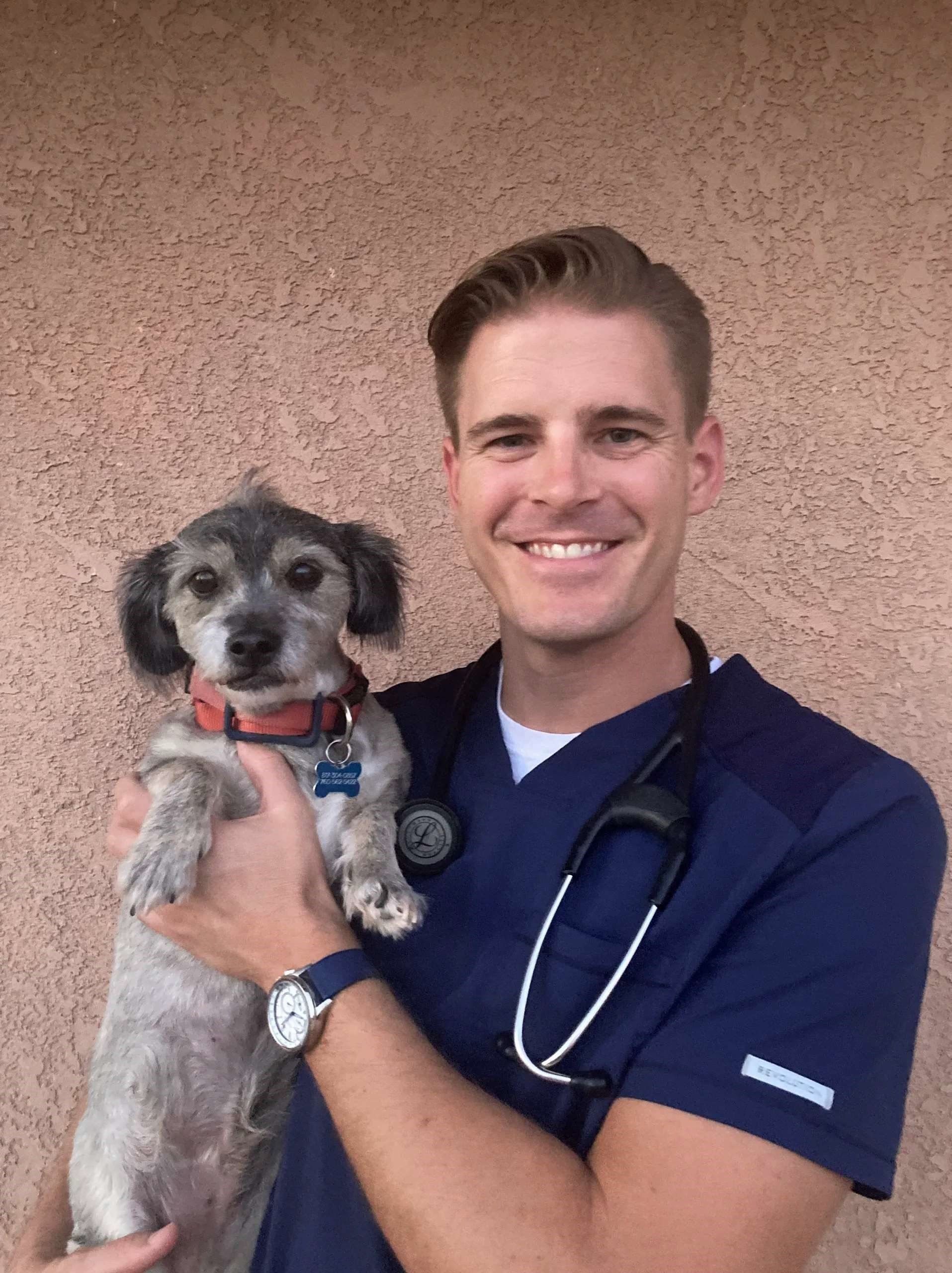 Man in scrubs holds small dog, both smiling. Stethoscope around his neck; background is textured wall. Man in scrubs holds small dog, both smiling. Stethoscope around his neck; background is textured wall.