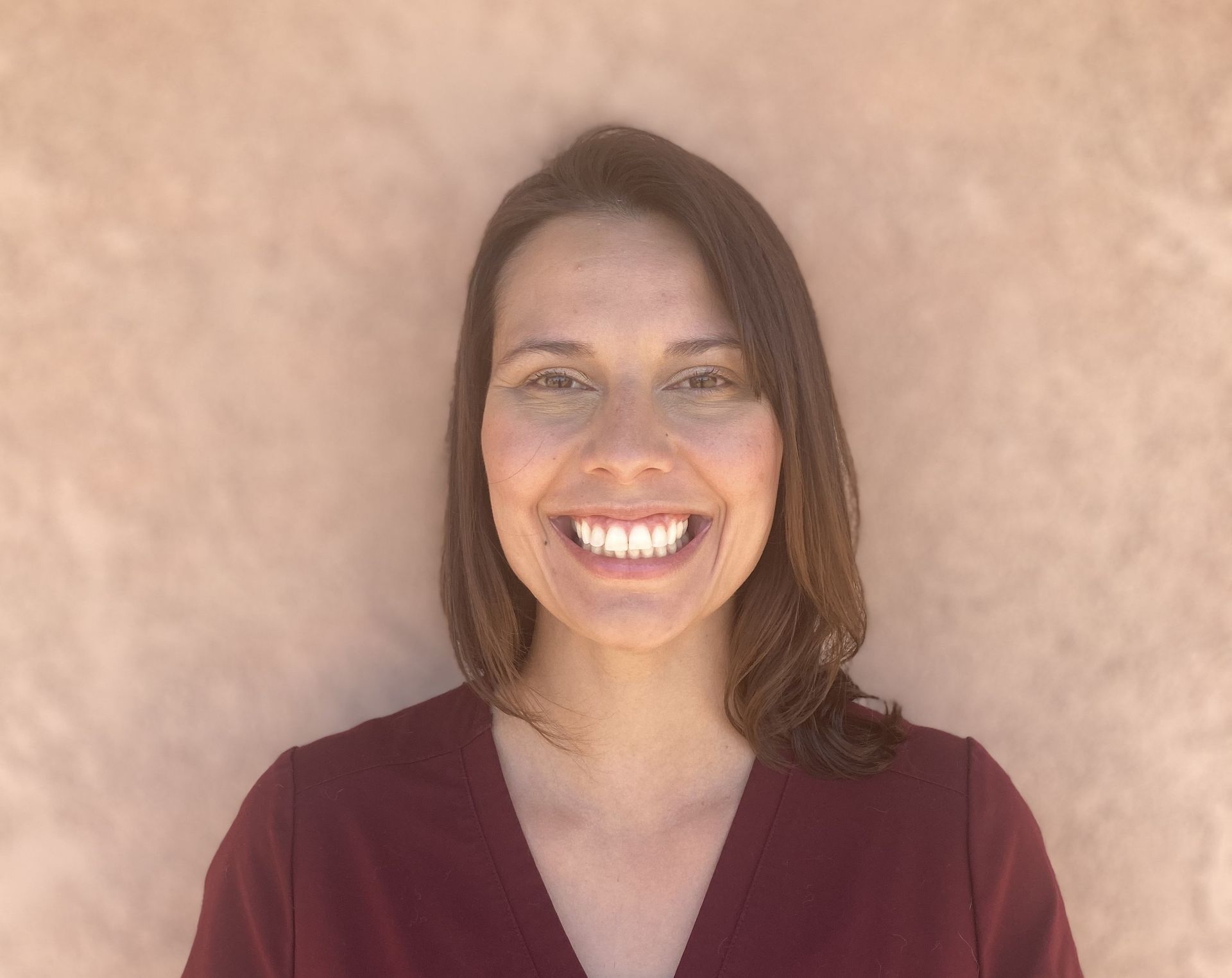 Woman with brown hair and a burgundy top smiles in front of a peach-colored wall. Woman with brown hair and a burgundy top smiles in front of a peach-colored wall.