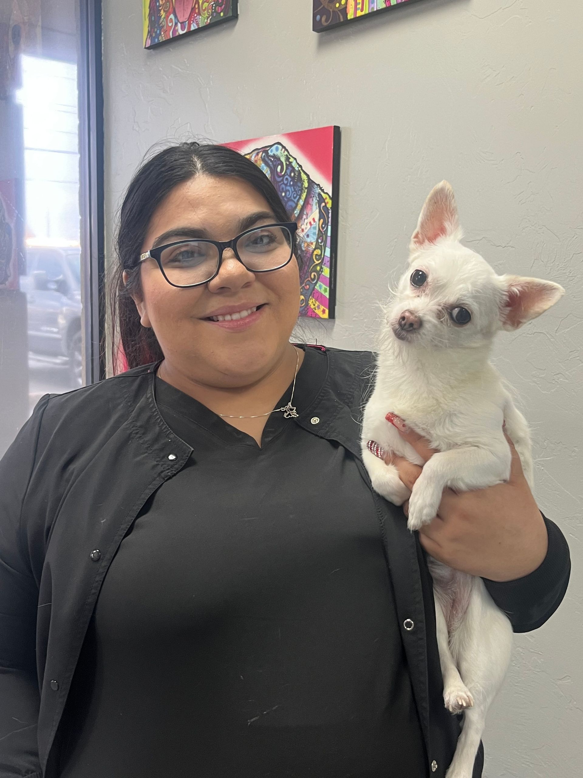 Woman in black shirt holding a small white dog, both smiling in front of a wall with artwork. Woman in black shirt holding a small white dog, both smiling in front of a wall with artwork.