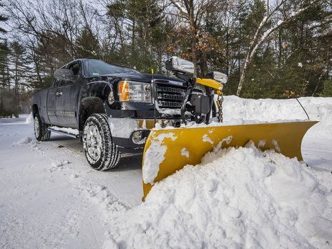 Black pickup truck with a mounted yellow snowplow blade clearing a heavy snowfall