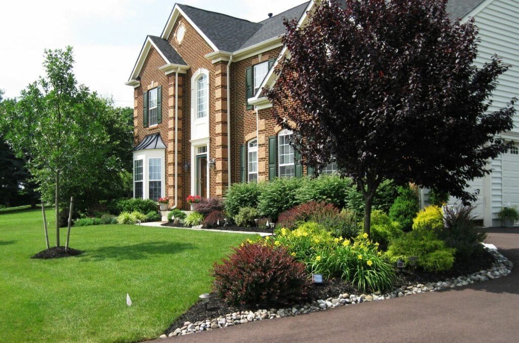 Manicured front lawn of a brick house with lush garden beds featuring a variety of shrubs and flowering plants, bordered by a curved path of river stones.