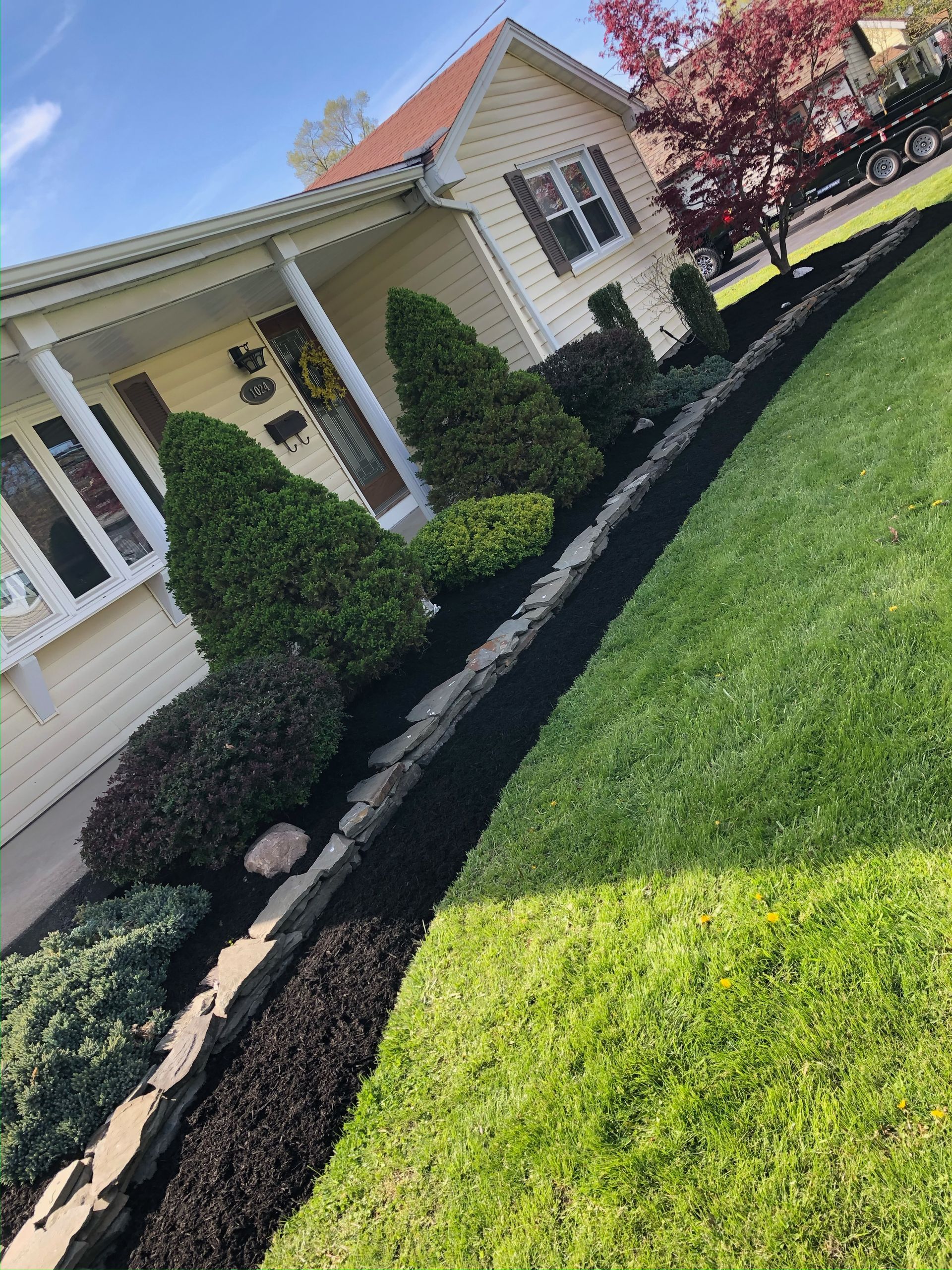 Landscape mulching in progress along a house's garden bed with conical shrubs and a slate stone border
