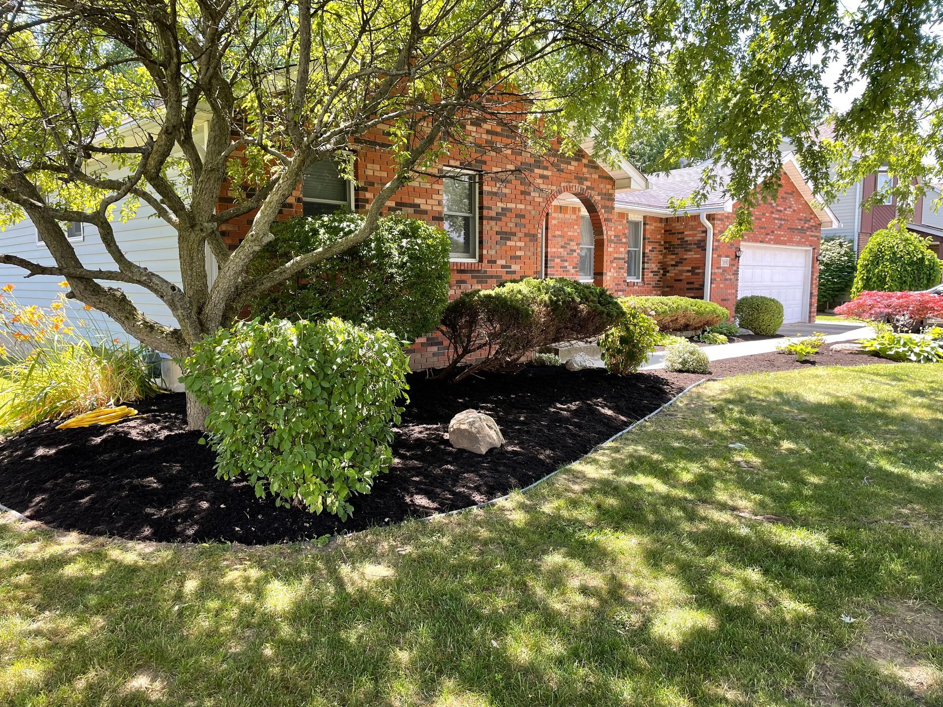 Well-maintained front yard of a brick house with mulched garden beds, shrubs, and a mature tree creating shade