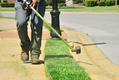 Worker trimming the edge of a neatly maintained lawn beside a paved sidewalk with a weed wacker