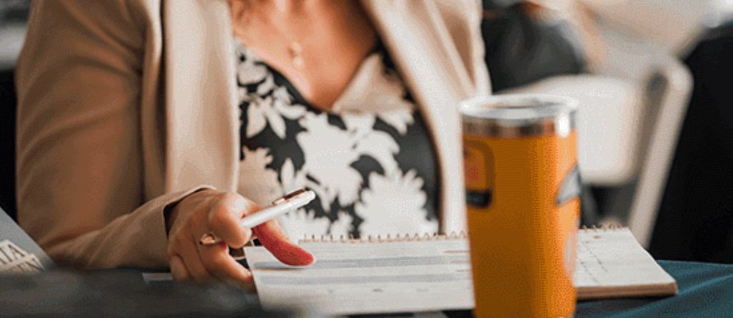A woman is sitting at a table holding a pen and a notebook.