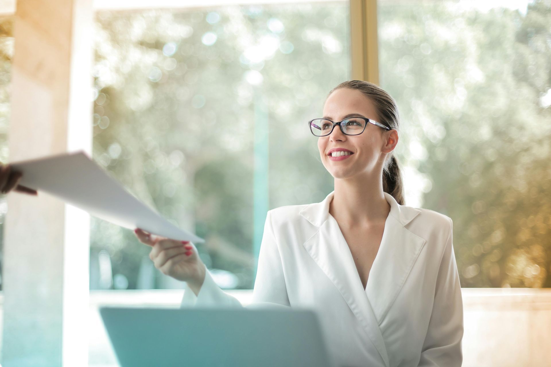 A woman is sitting at a desk with a laptop and holding a piece of paper.