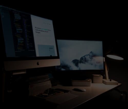 Two computer monitors are sitting on a desk in a dark room.