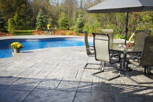 Patio with glass table, chairs, and umbrella beside a blue swimming pool and wooded yard