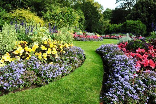 Colorful flower beds border a curving green garden path with trees and blooming shrubs.