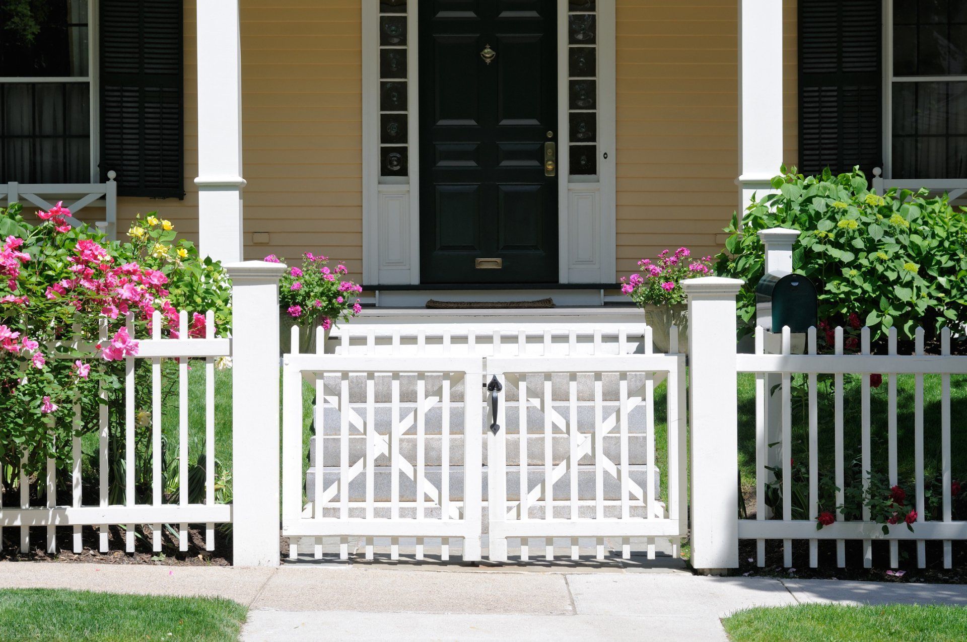 White picket gate and fence in front of a house with pink flowers and green shrubs