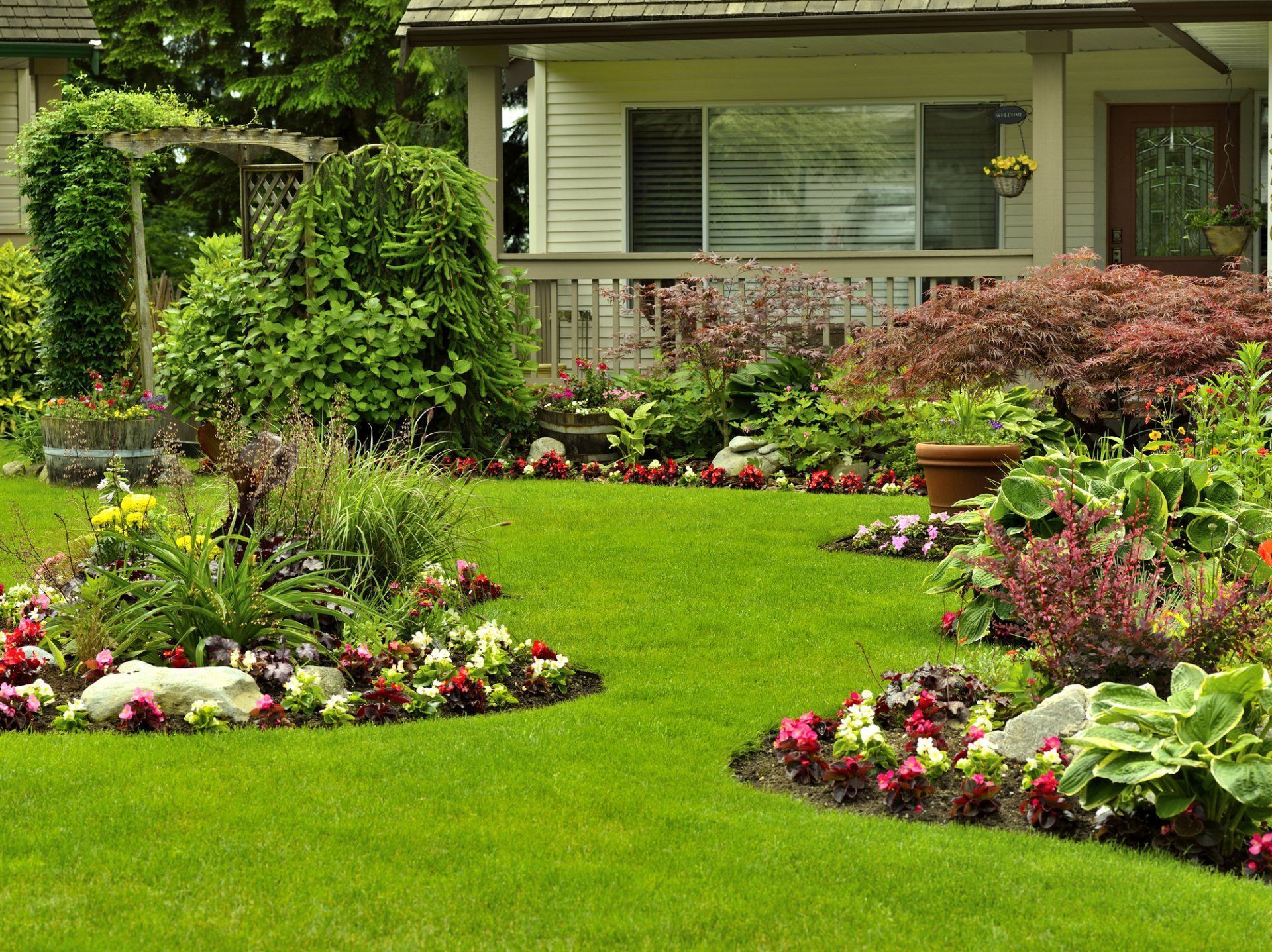Front yard with lush green lawn, colorful flower beds, and a house porch in the background