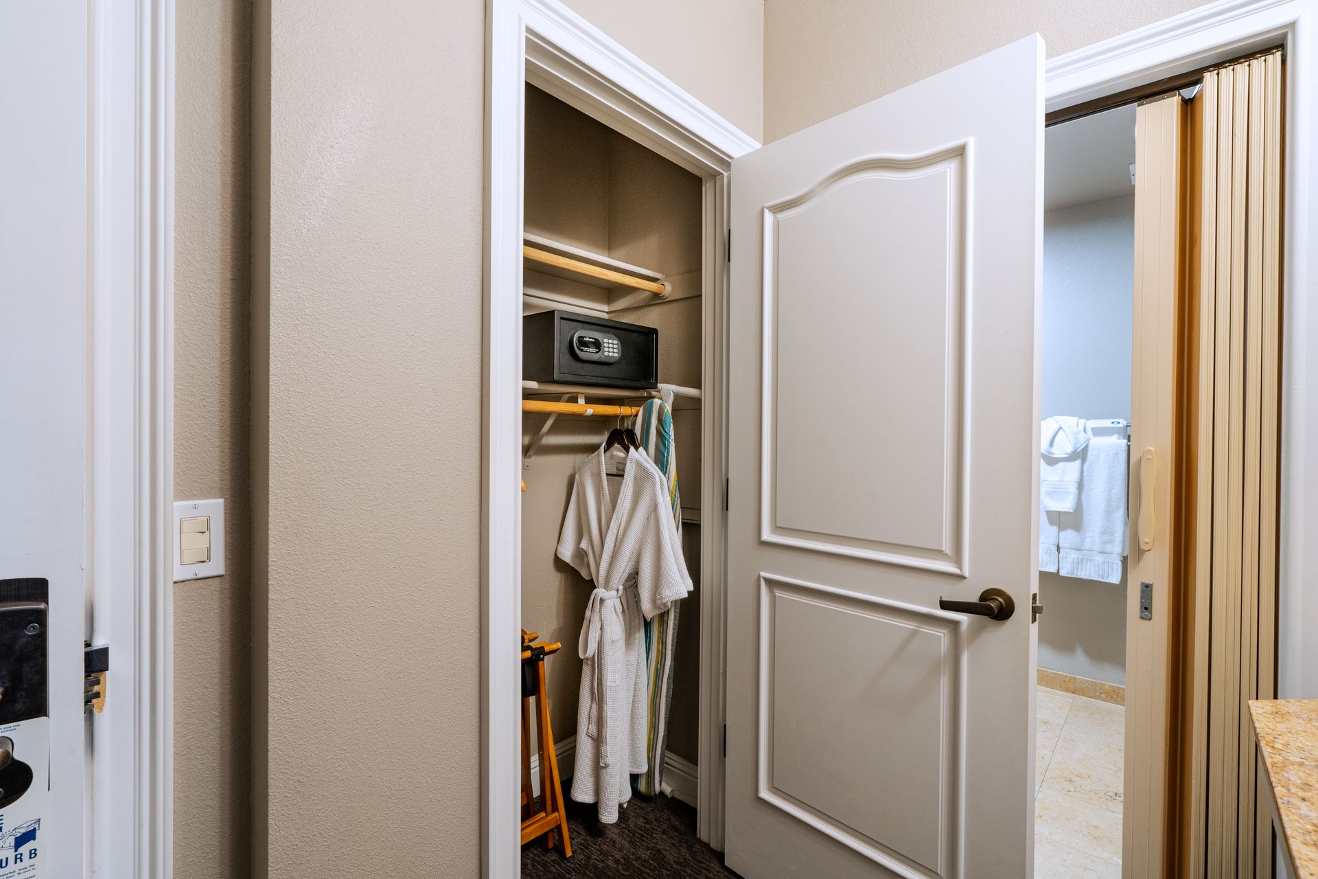 Closet with robe, safe, and open door to bathroom with towels. Light walls and door.