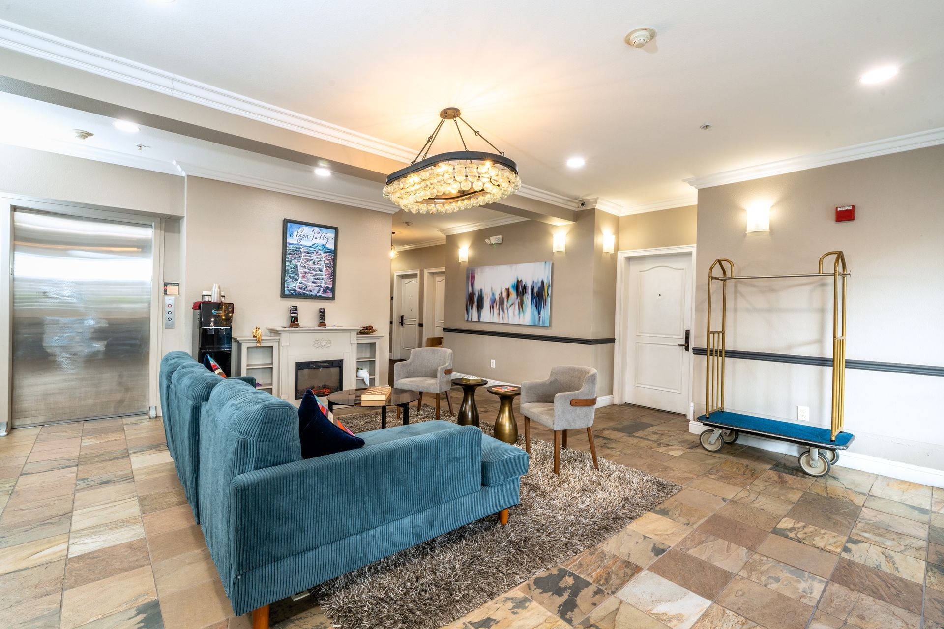 Hotel lobby with a teal sofa, chairs, and chandelier. Includes an elevator, luggage cart, and artwork.