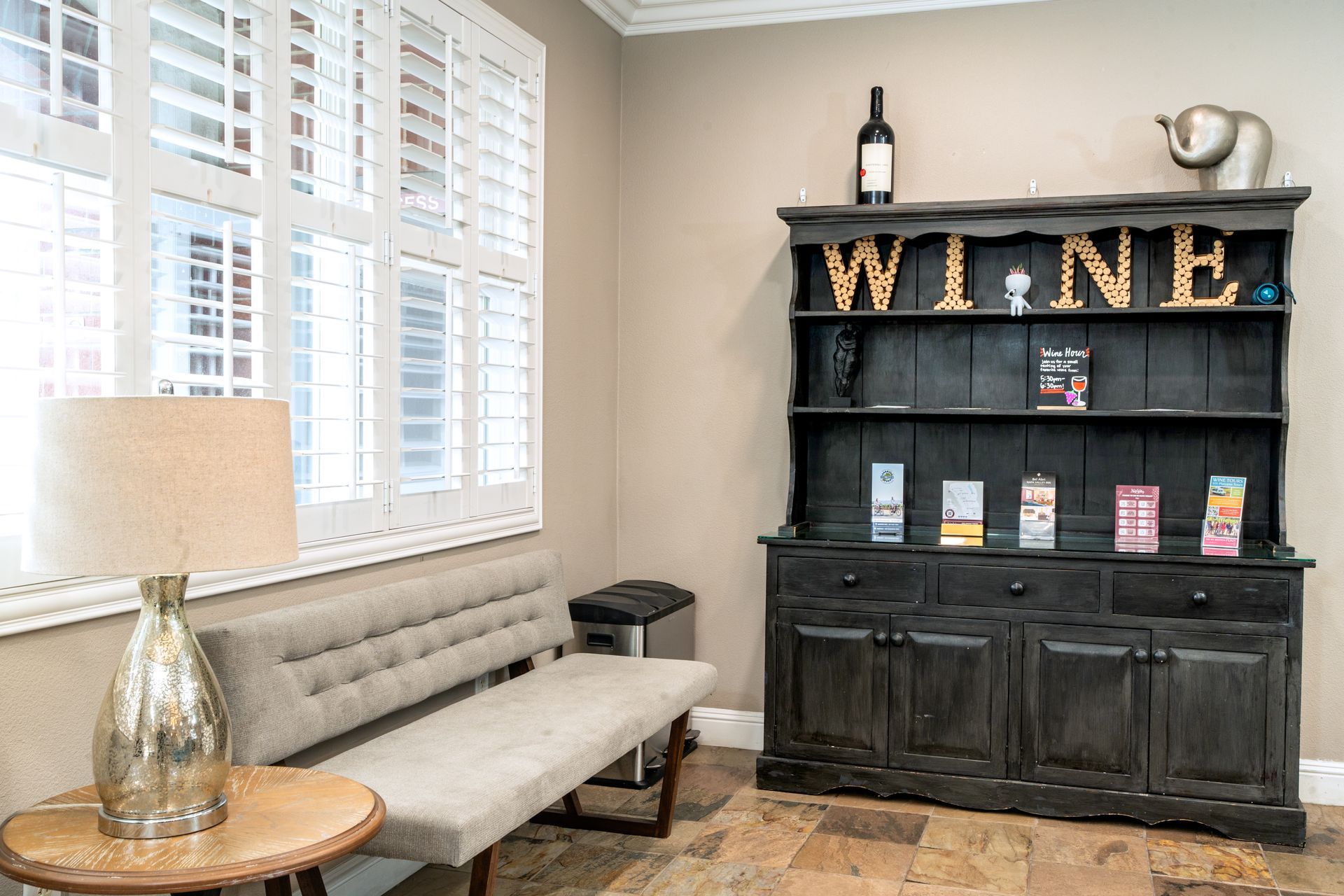 A waiting area with a bench, lamp, and wine-themed display cabinet next to a window with shutters.