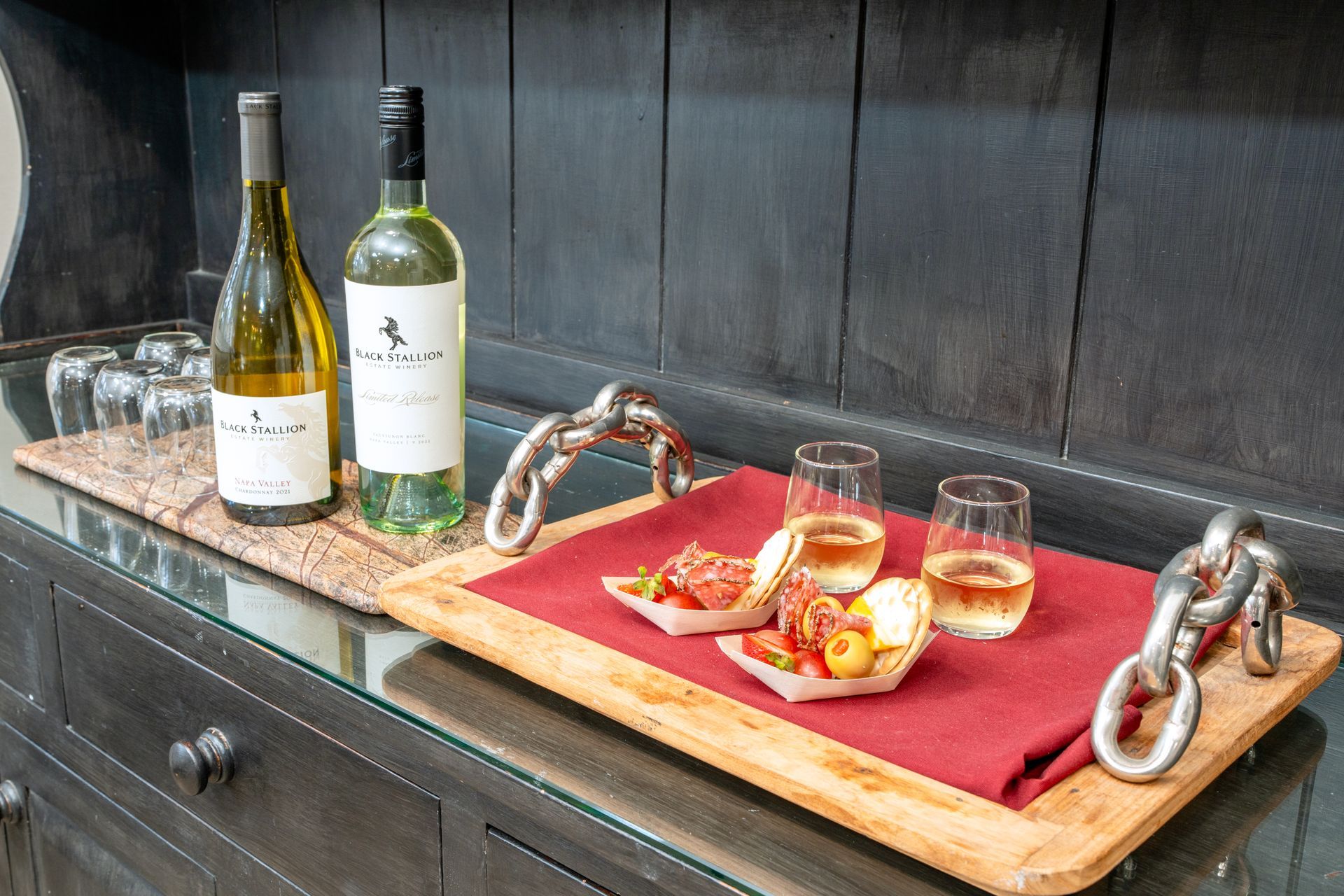 Wine bottles, charcuterie, and wine glasses on a wooden tray atop a dark cabinet.