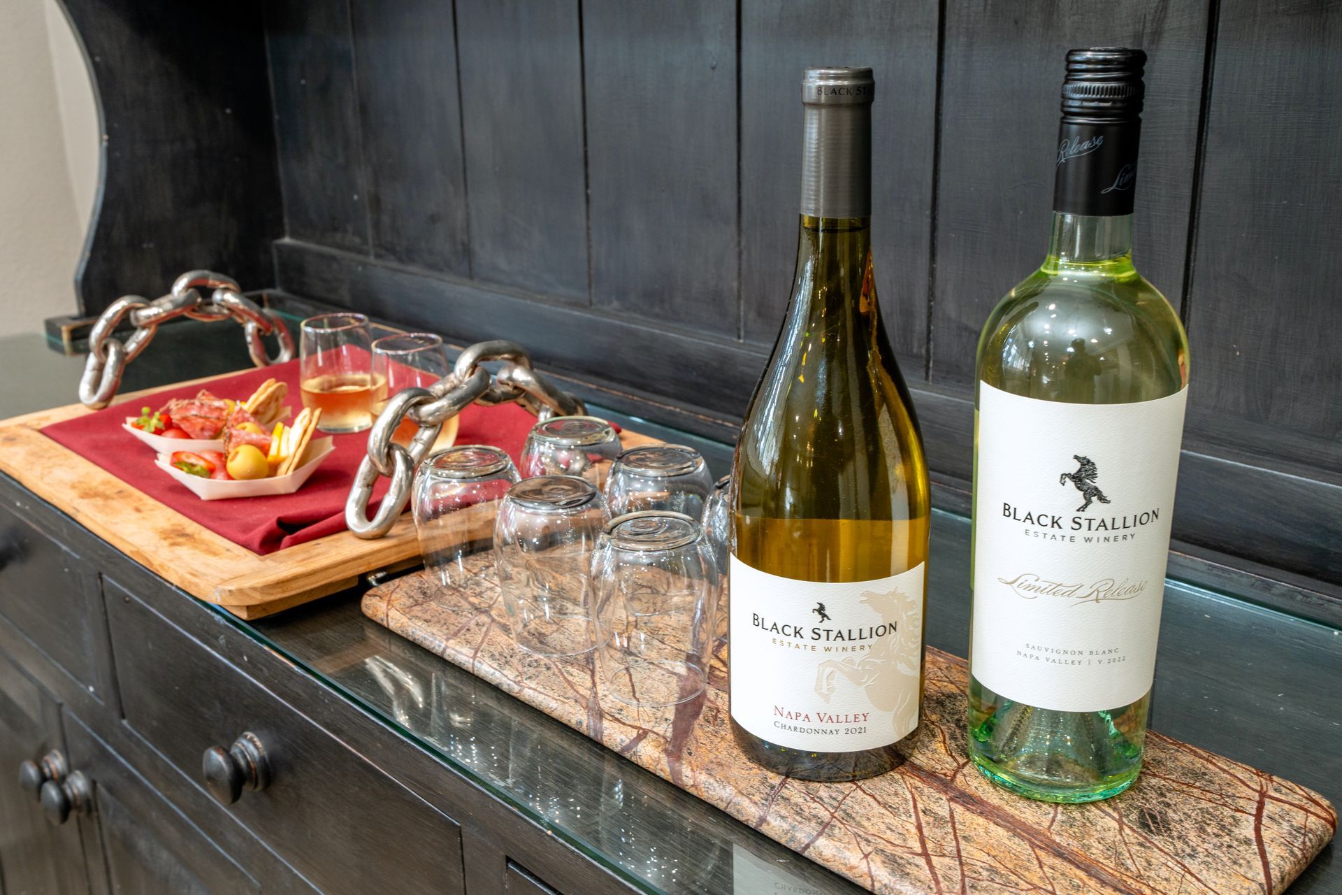 Two wine bottles, glasses, and snacks on a dark wooden cabinet, ready for serving.