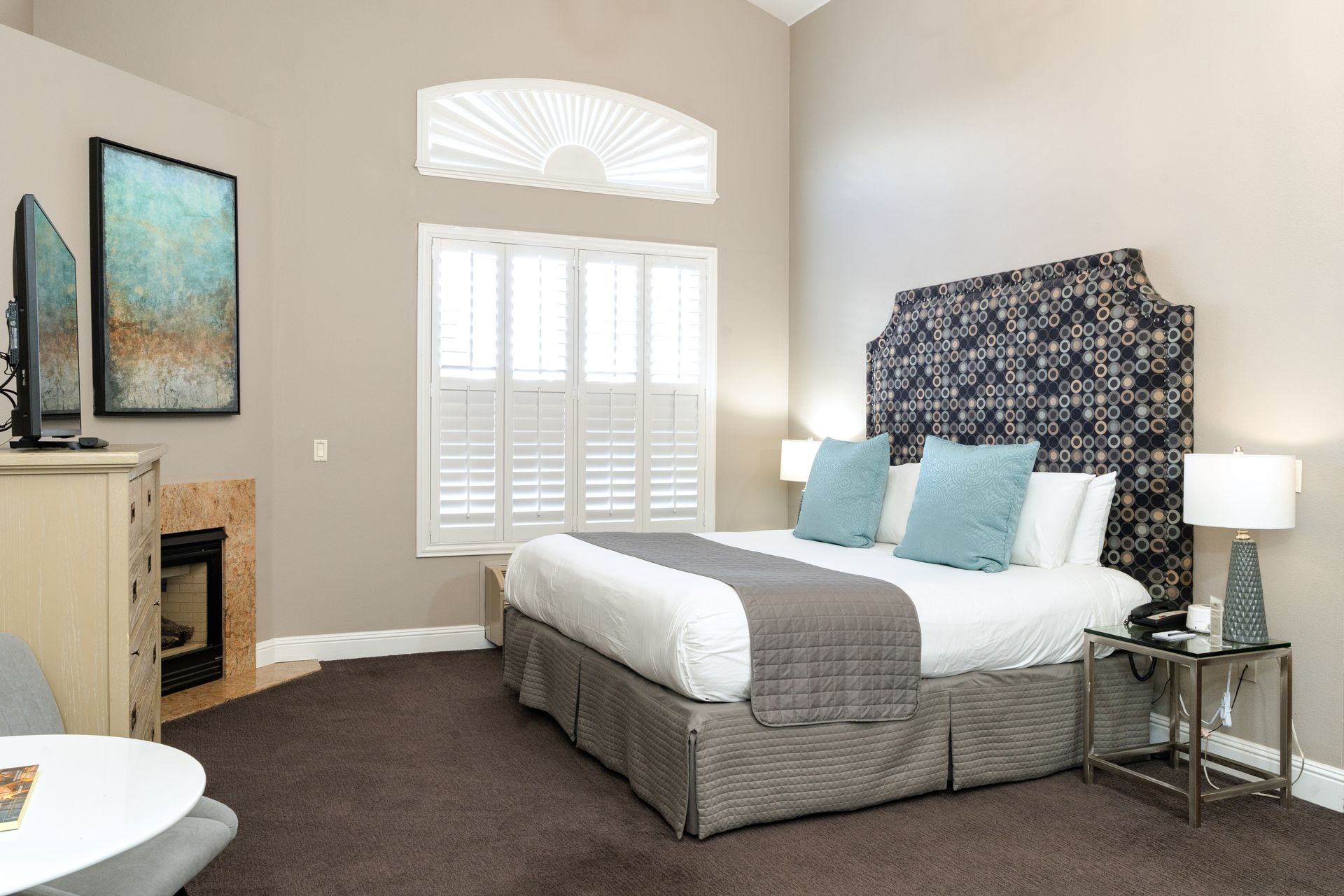 Bedroom with bed, headboard, fireplace, artwork, and window with shutters.