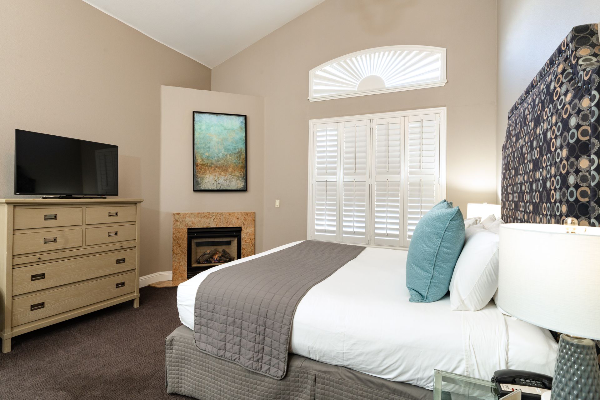 Bedroom with bed, fireplace, TV, and shuttered window. Earth-toned walls and blue accents.