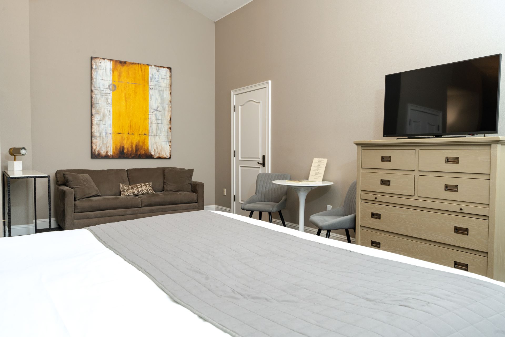 Bedroom with a bed, sofa, dresser, table, chairs, and artwork. Neutral tones with a pop of yellow.
