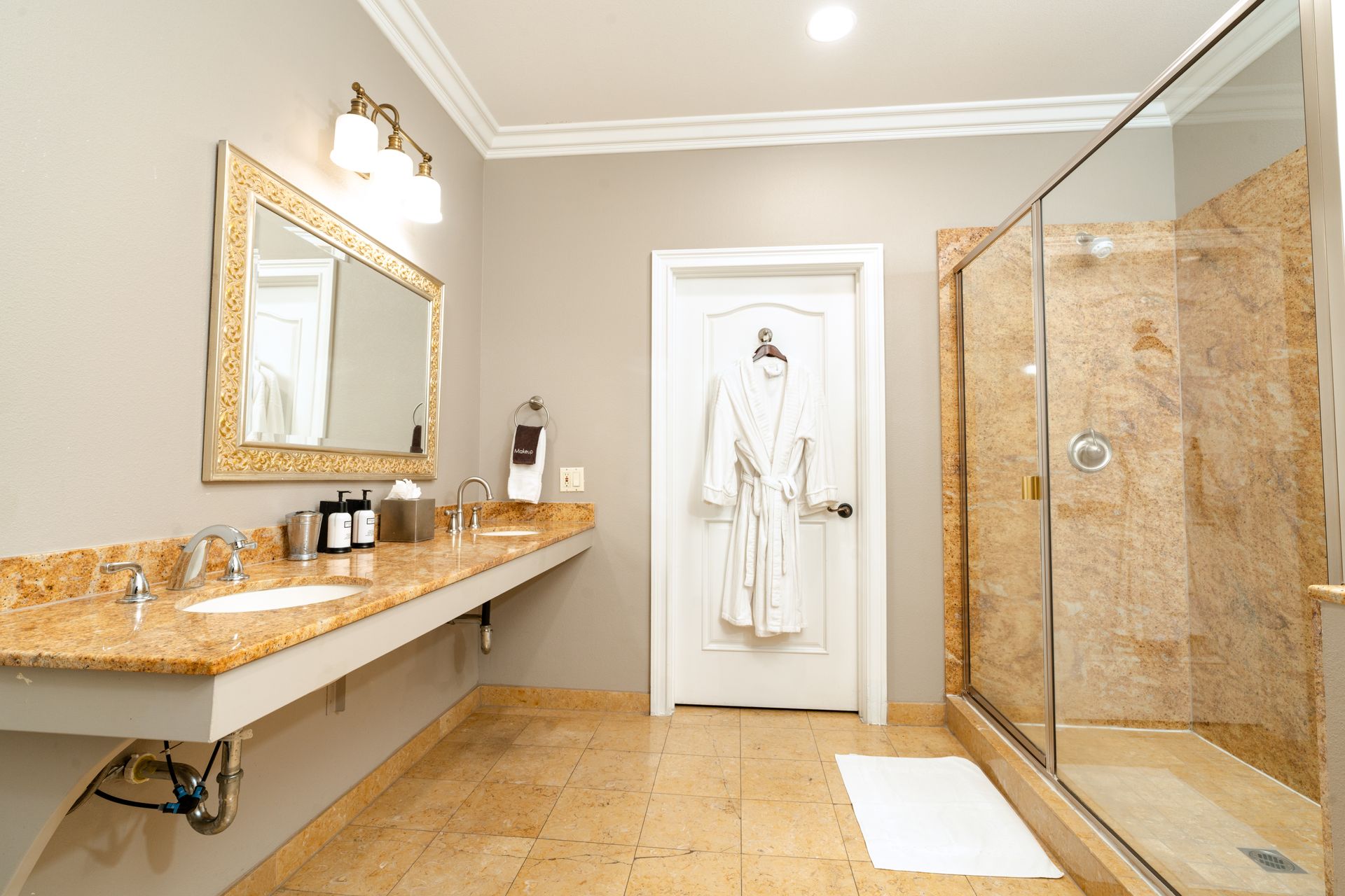 Spacious bathroom with beige walls, granite countertop, glass shower, and white bathrobe hanging on the door.