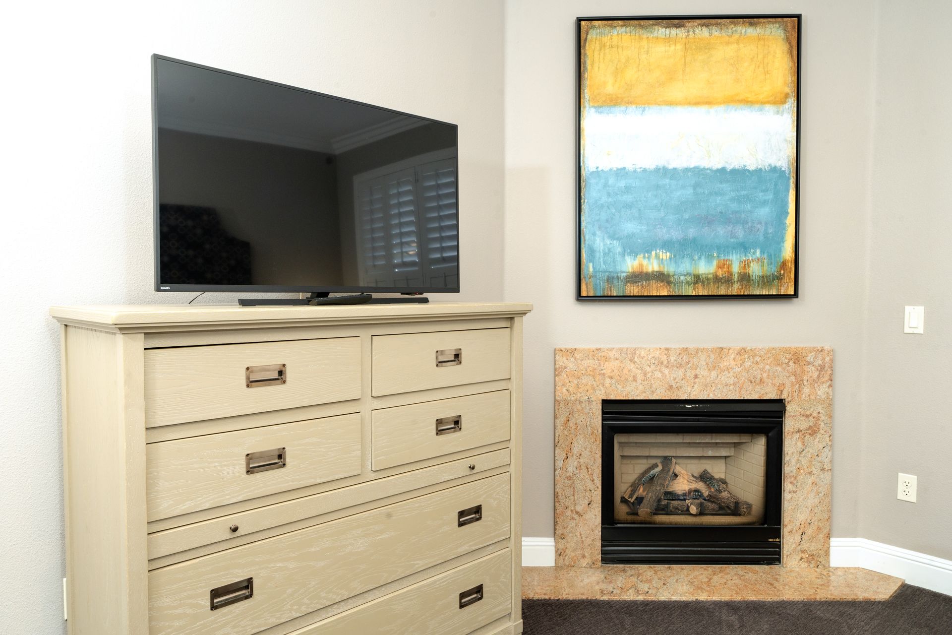 A TV on a beige dresser next to a fireplace with a painting above it in a corner of a room.