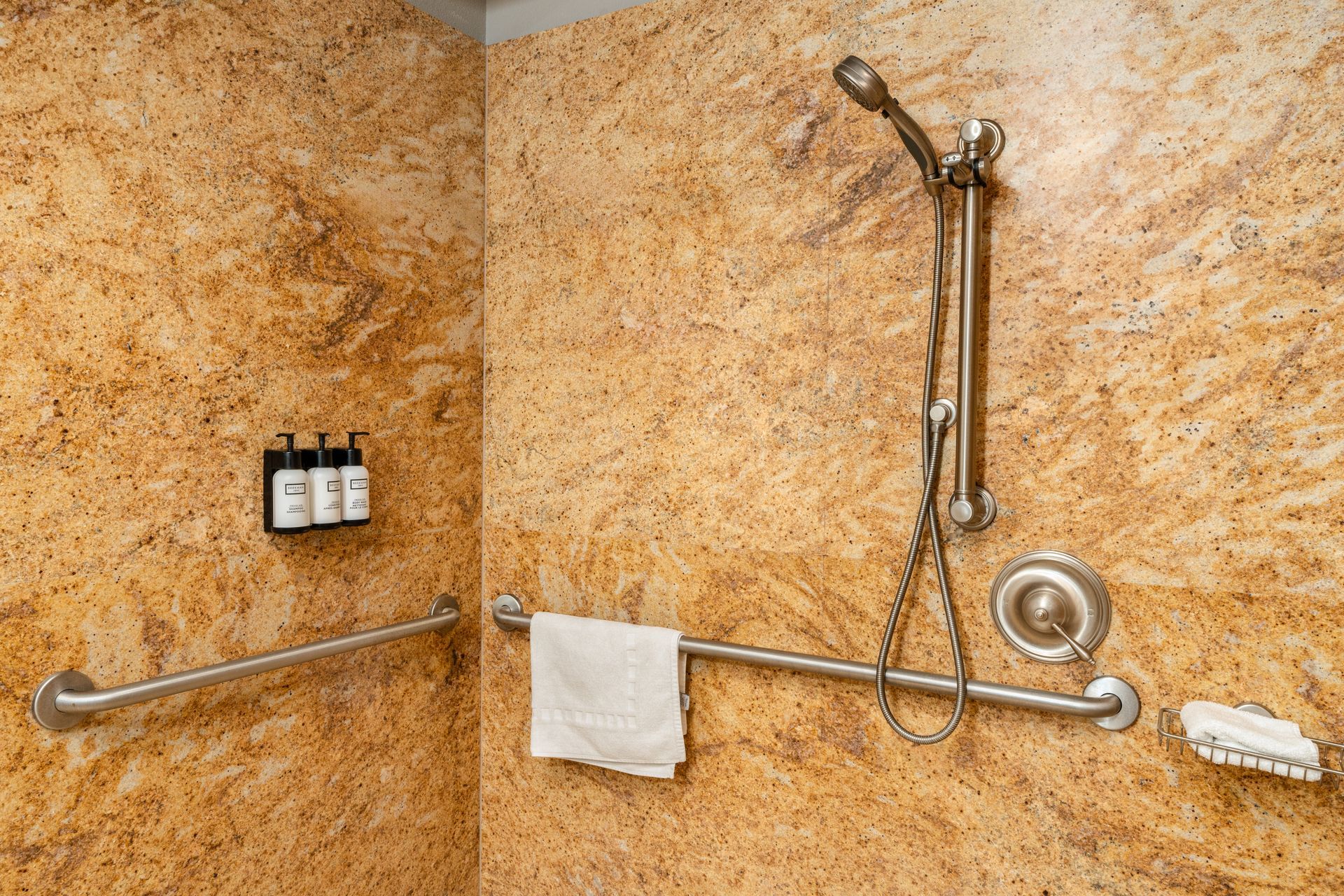 Shower stall with granite-like walls. Handheld shower head, grab bar, soap dish, and toiletries are visible.