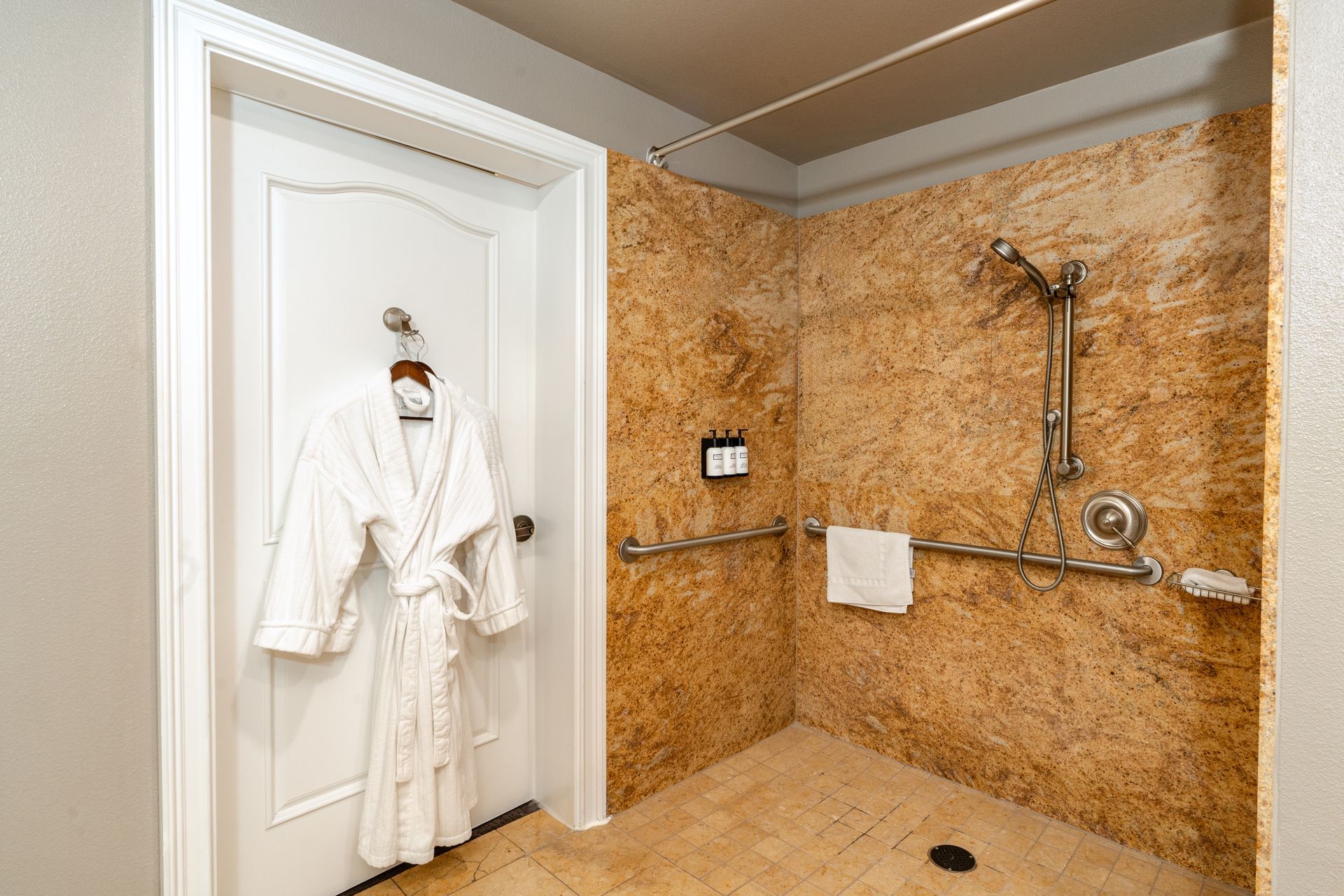 Accessible bathroom with walk-in shower. Beige walls, safety bar, robe on door.