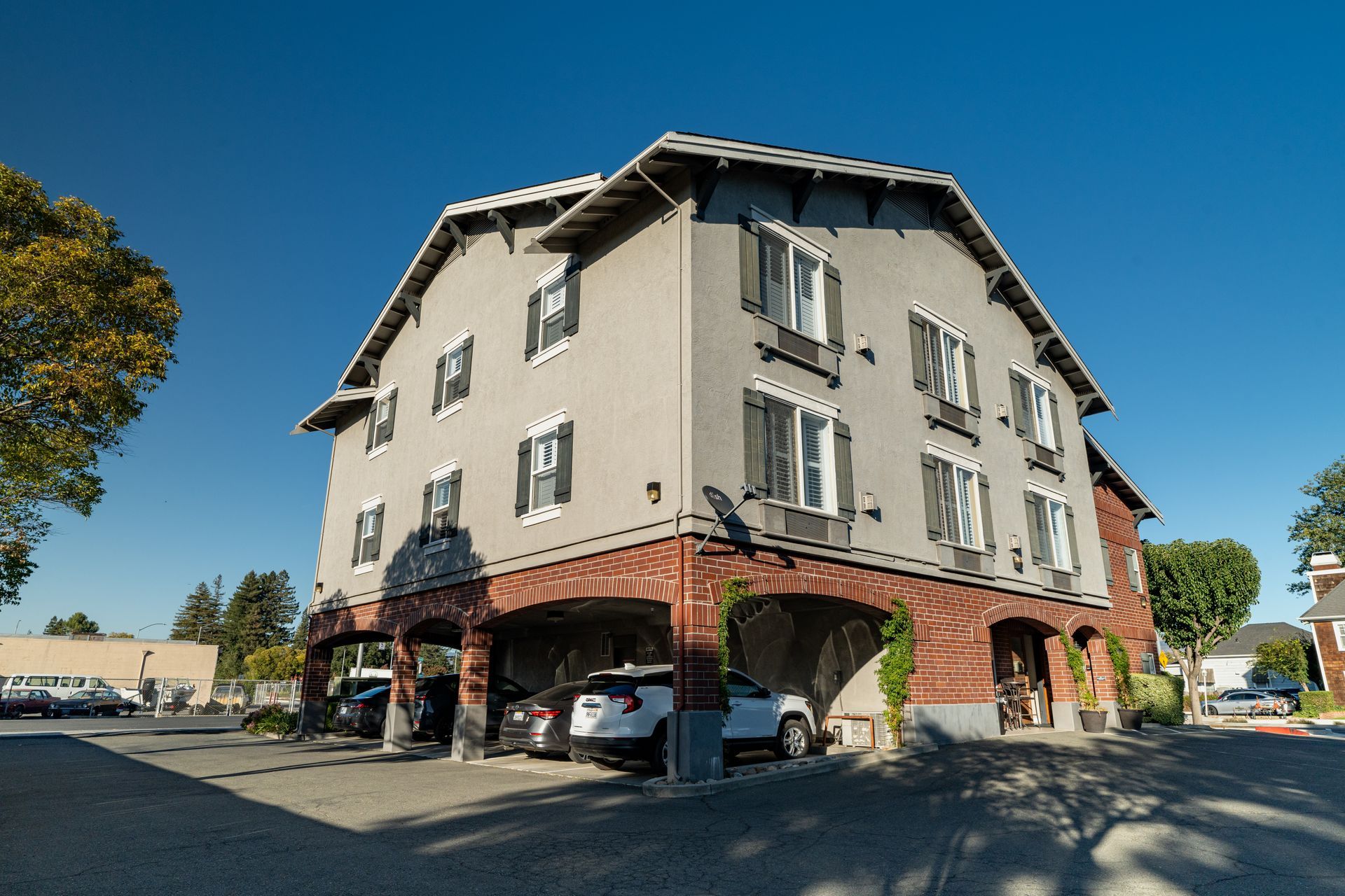 Three-story gray building with red brick arches, parked cars underneath, against a clear blue sky.