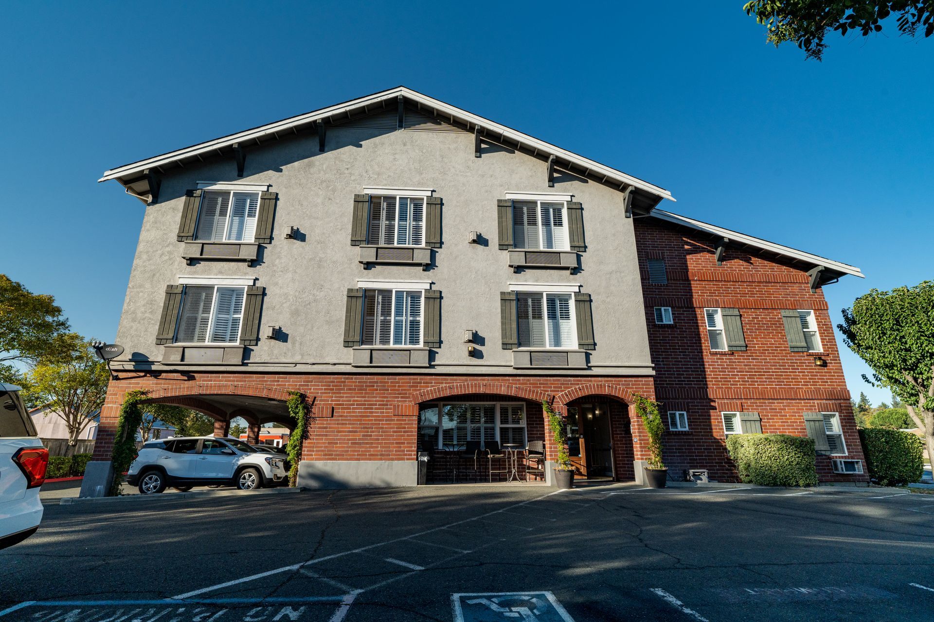 Three-story building with gray stucco and red brick facade, white shutters, arched entrance, and car parked beneath.