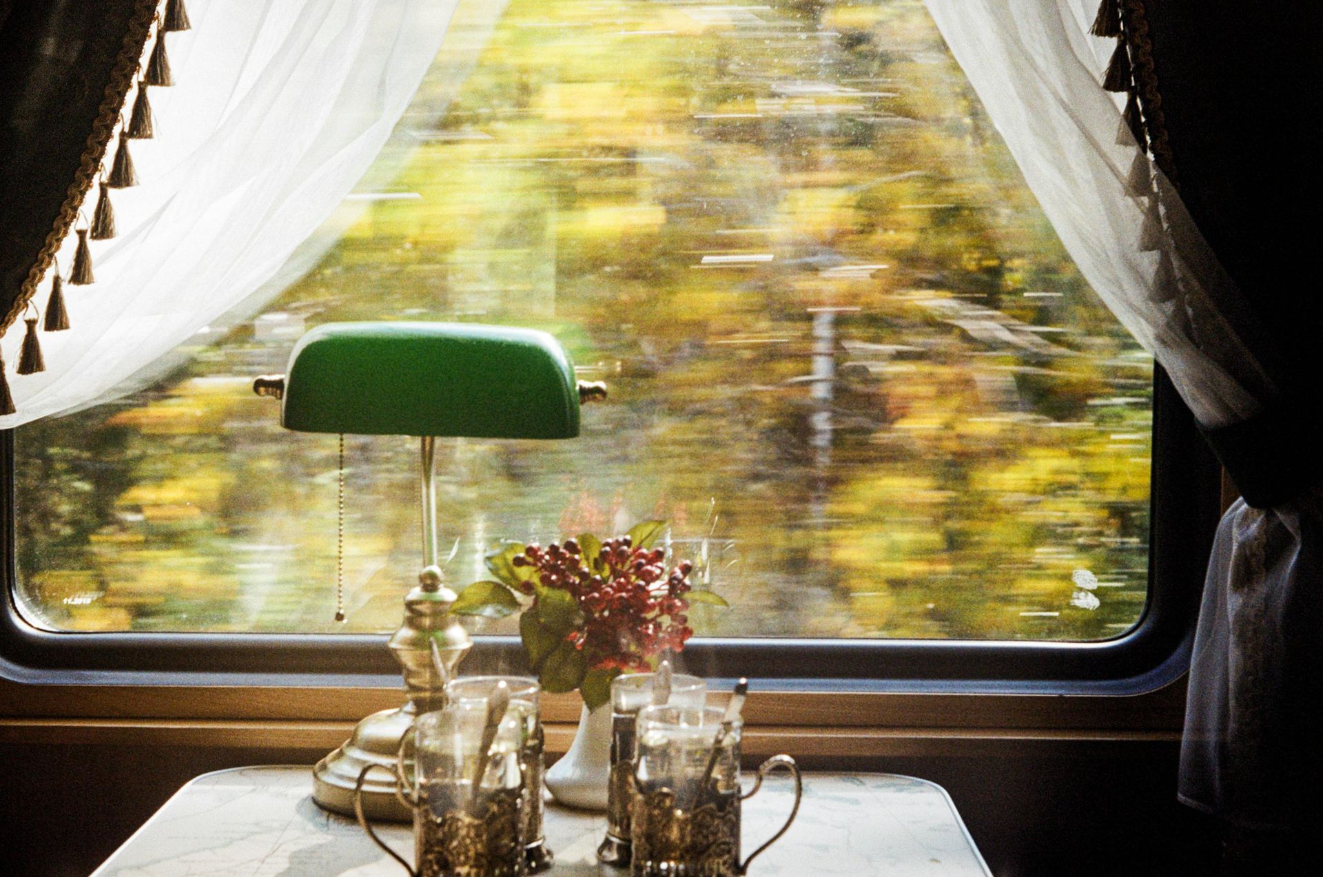 Train interior with a view of blurred trees, a table with glassware and a lamp.
