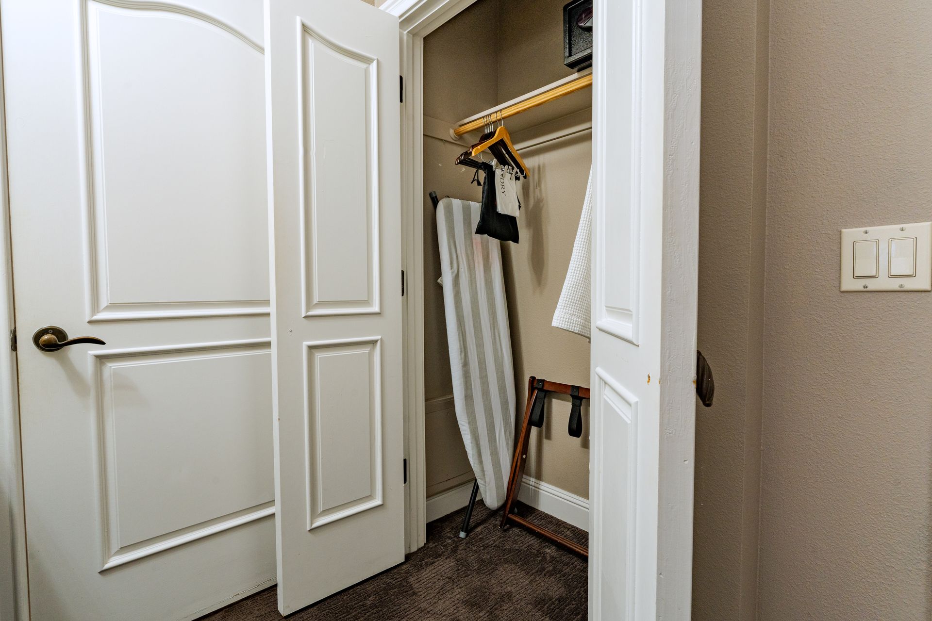 Open closet with an ironing board, hangers, and shelf. White doors and tan walls.