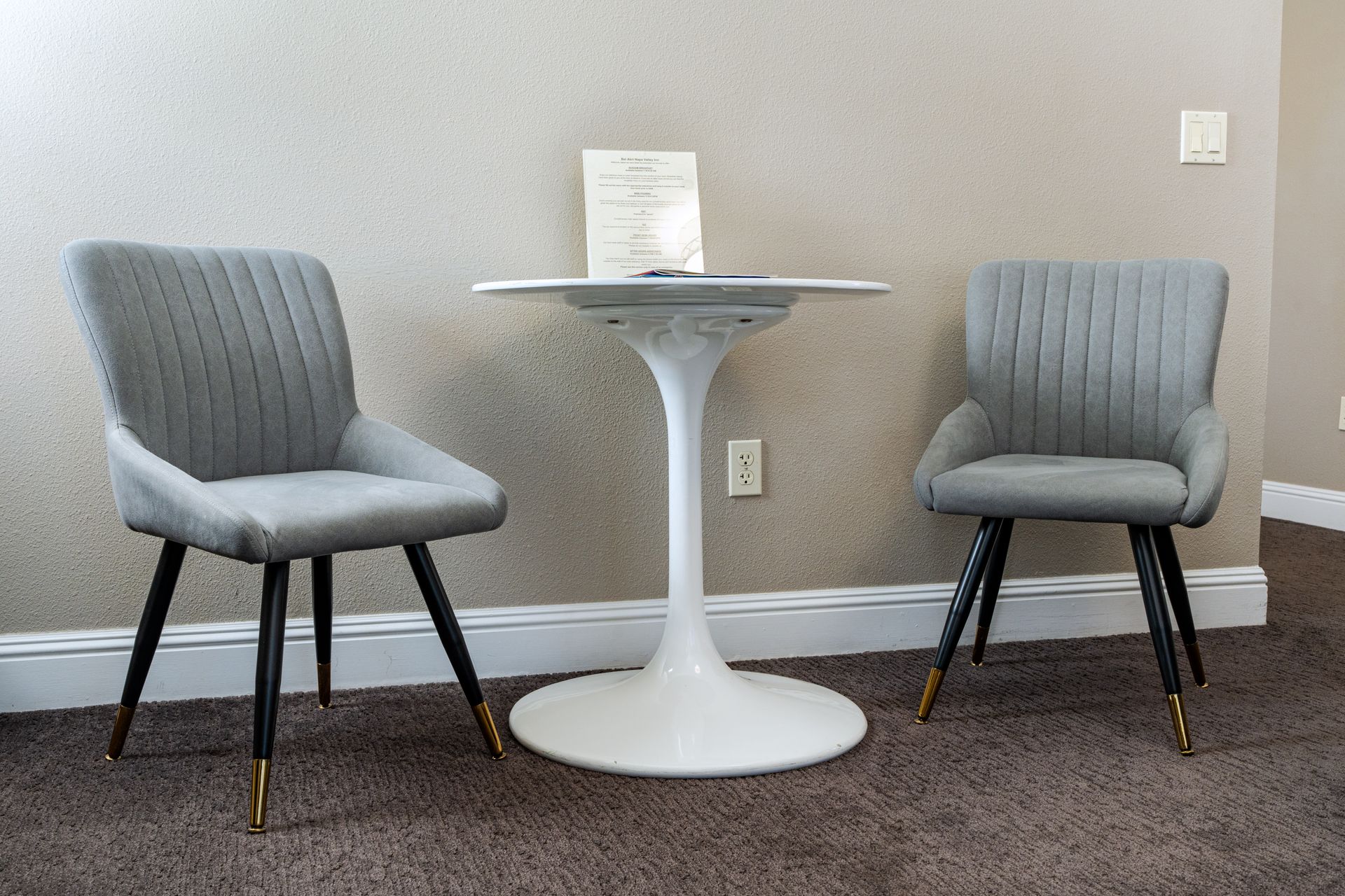 Two gray upholstered chairs and white tulip table on brown carpet against beige wall.