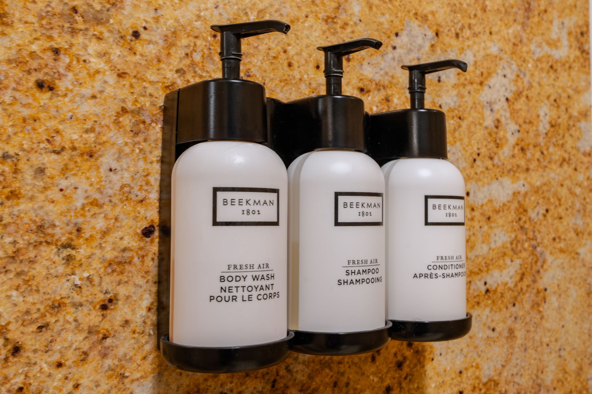 Three white bottles with black pumps in a wall-mounted holder on a textured, brown surface.