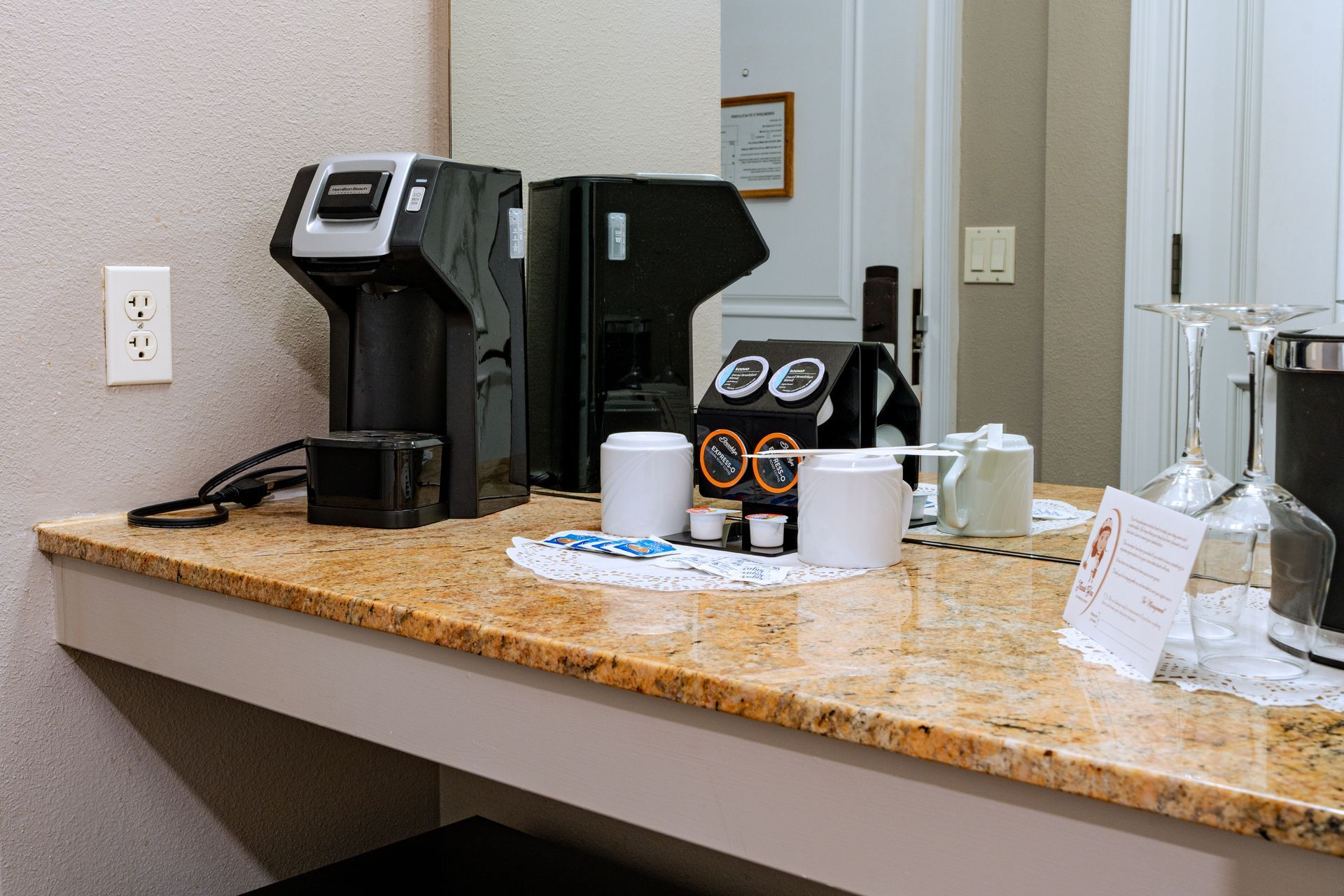 Coffee station on a countertop with a coffee maker, cups, and condiments. Mirror reflects the room.