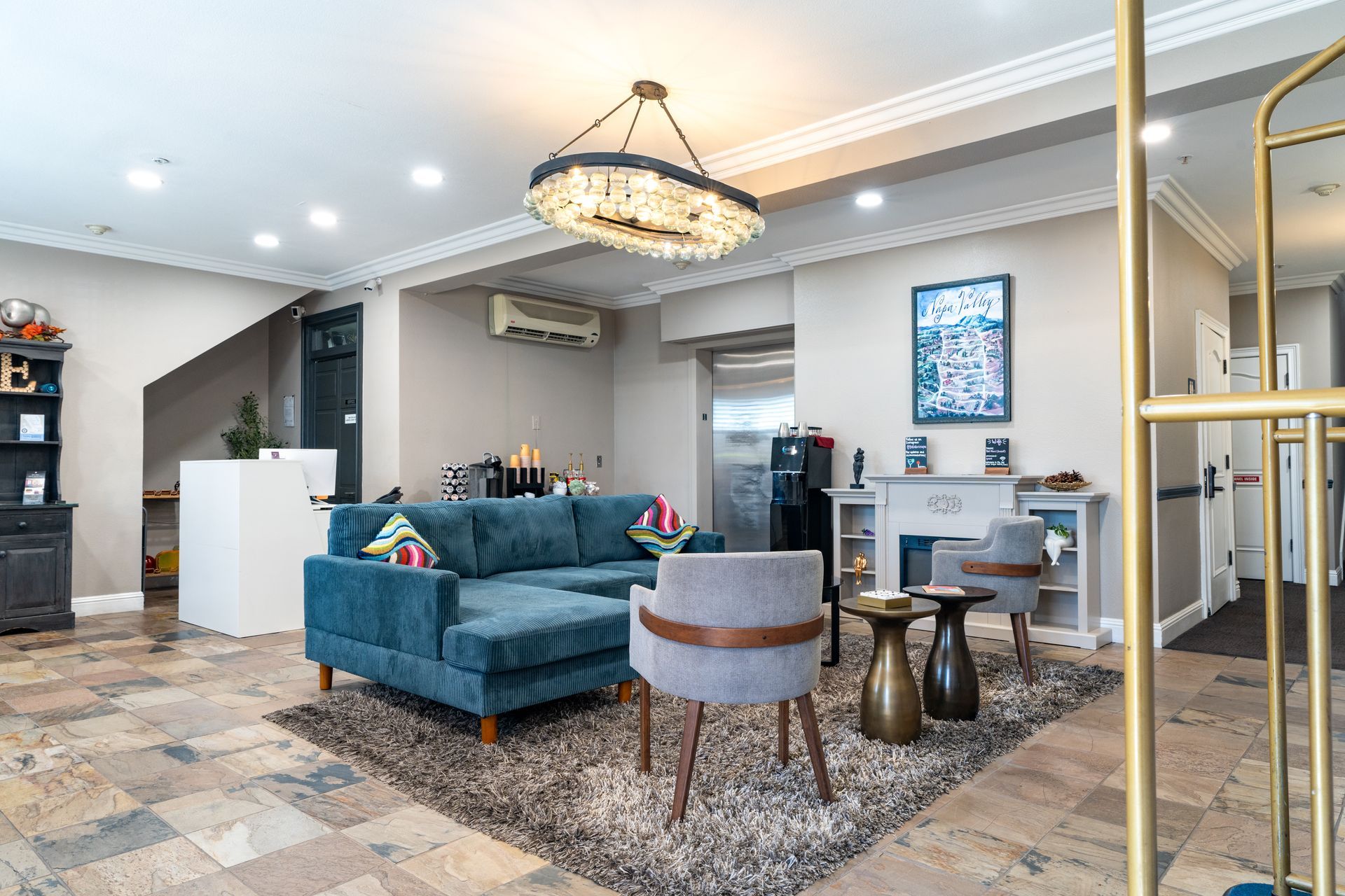 Lobby with blue sofa, two gray chairs, coffee table, and luggage cart, in front of a reception desk and decorative art.
