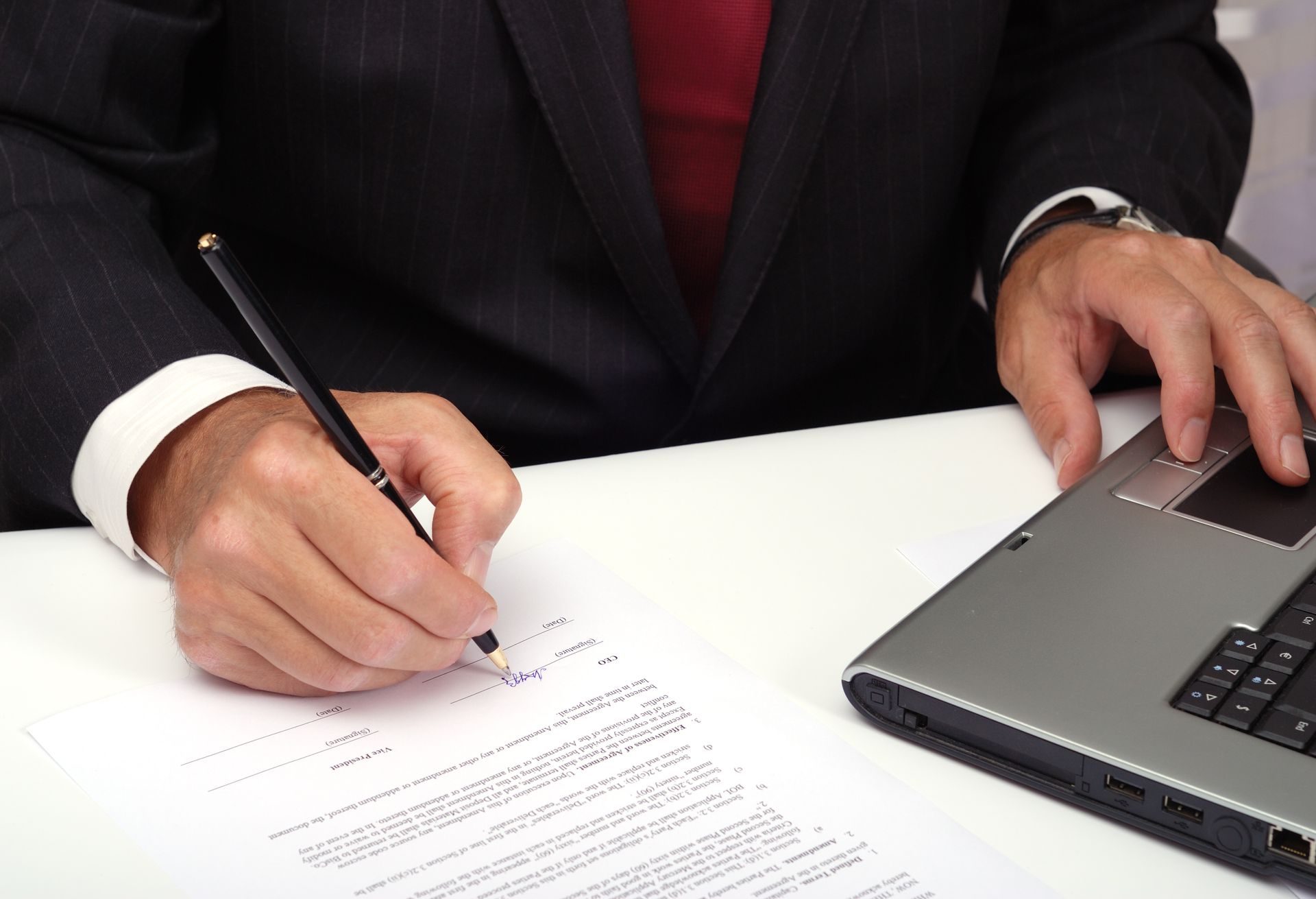 A man in a suit is writing on a piece of paper next to a laptop