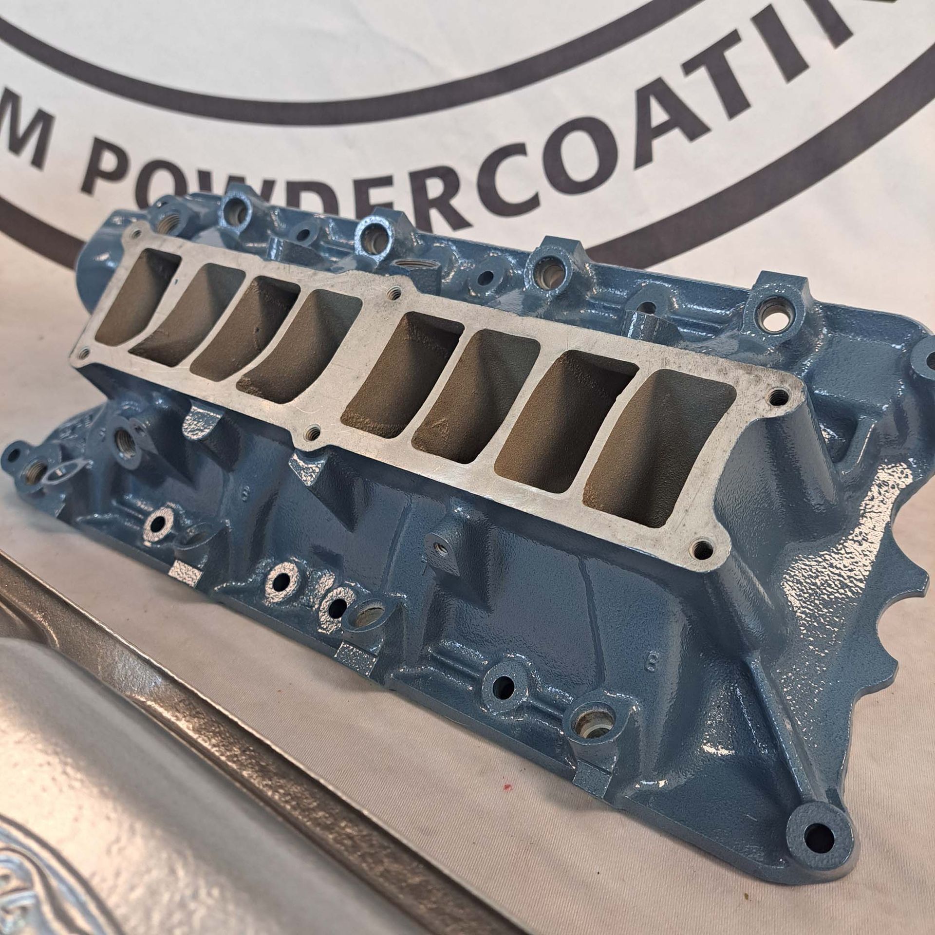 A blue engine intake manifold sits on a table in front of a sign that says powdercoat