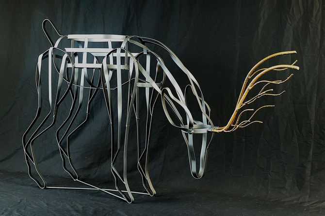 Metal sculpture of a deer, silver body with dark legs, drinking water, with golden antlers, set against a dark backdrop.