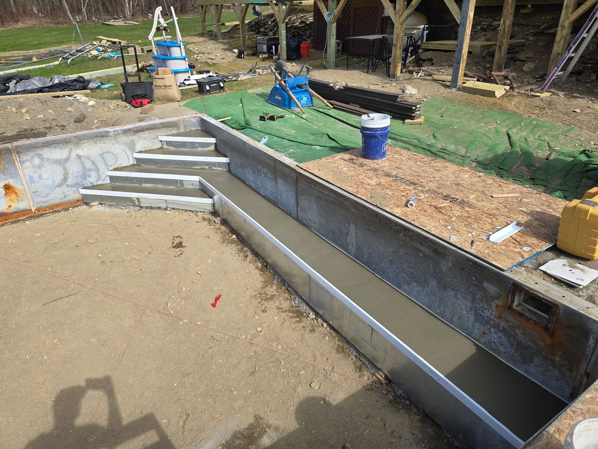 A bucket of concrete is sitting on the ground next to a swimming pool under construction.