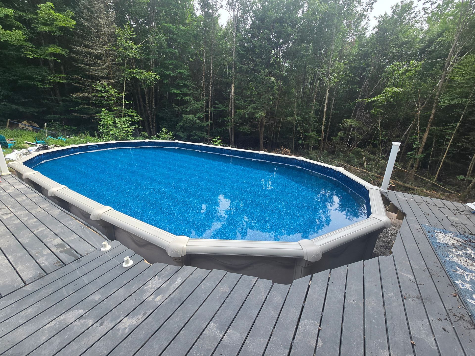 An oval swimming pool is sitting on top of a wooden deck surrounded by trees.