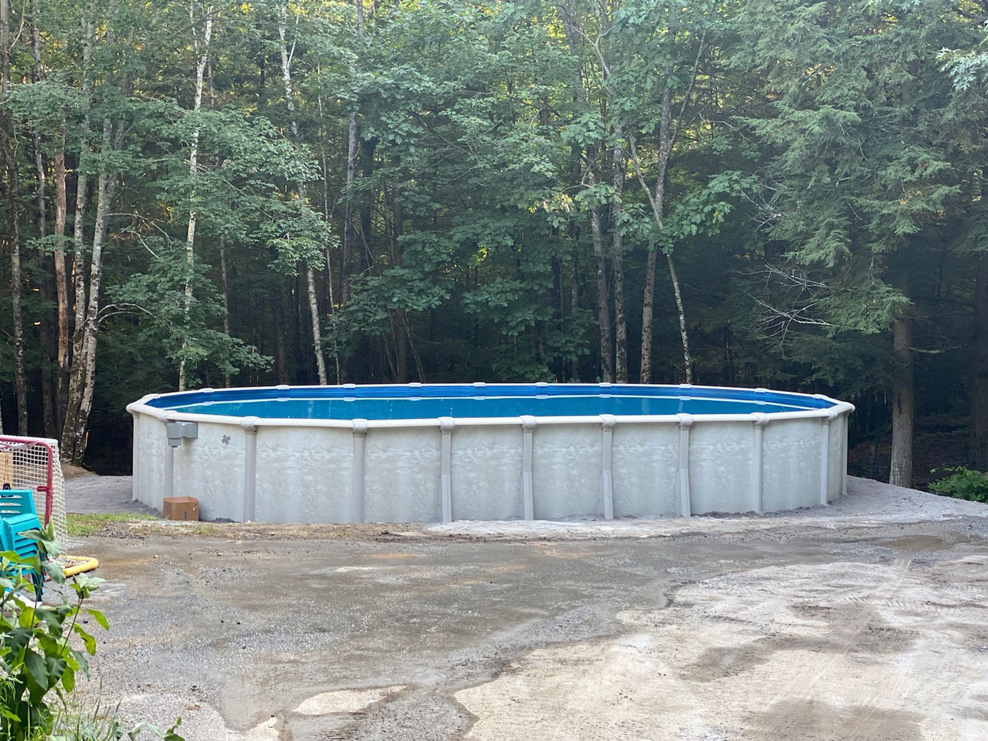 A large swimming pool is sitting in the middle of a dirt field surrounded by trees.