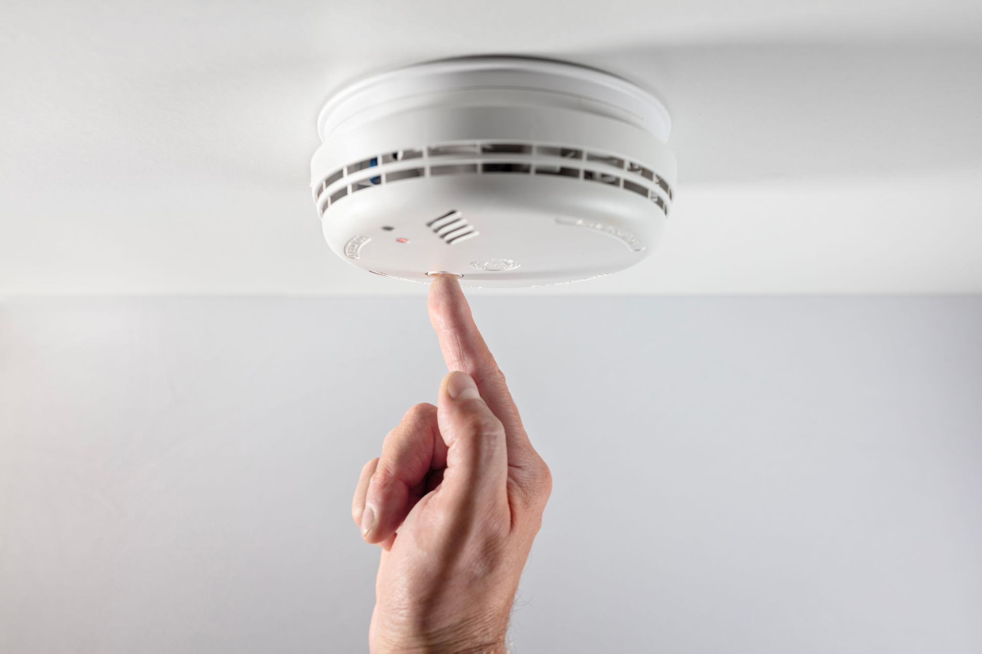 Hand testing a white smoke detector mounted on a white ceiling.