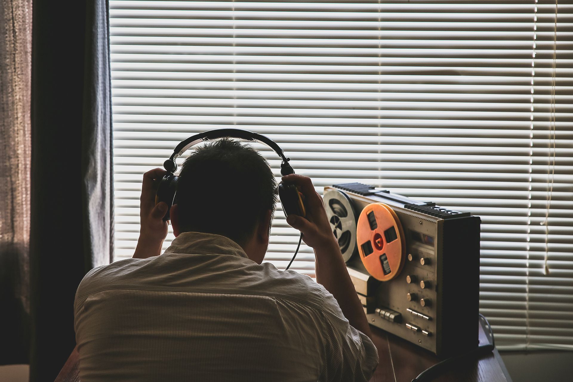 Man wearing headphones, listening to reel-to-reel tape recorder near a window with closed blinds.